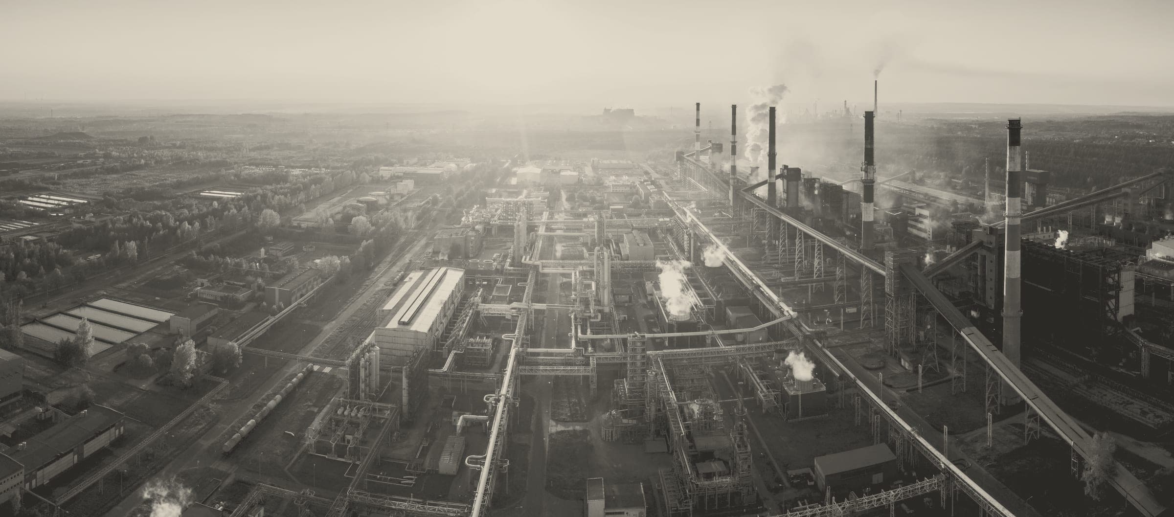 A wide sepia-toned aerial panorama of a sprawling heavy industrial complex, likely a petrochemical or steel plant, seen from an elevated vantage point. Tall chimneys emit dark plumes of smoke into a h