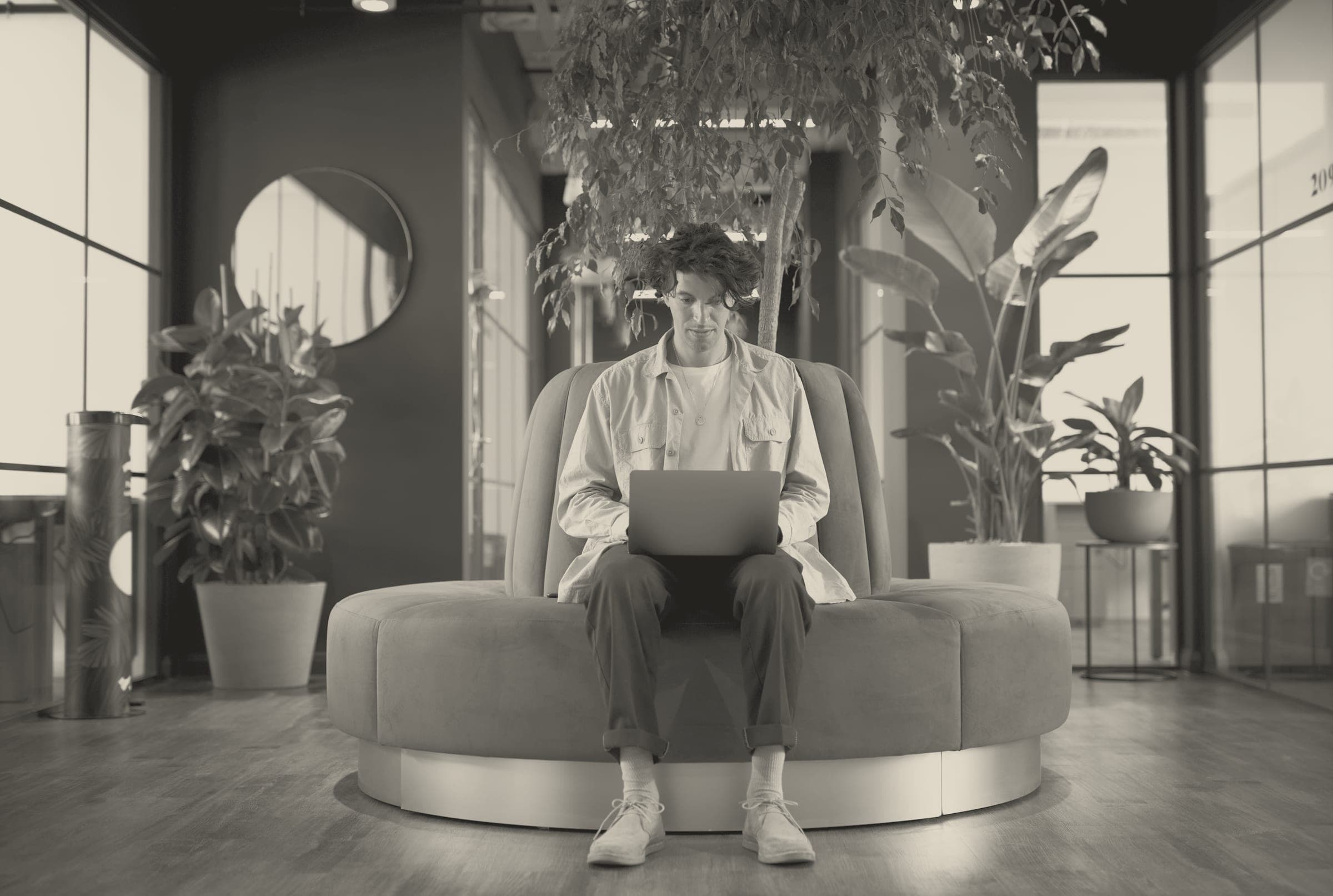 A young man with curly hair sits centrally on a large round upholstered lounge sofa in a modern office or hotel lobby, working on a laptop balanced on his knees. He wears a light overshirt, T-shirt, d